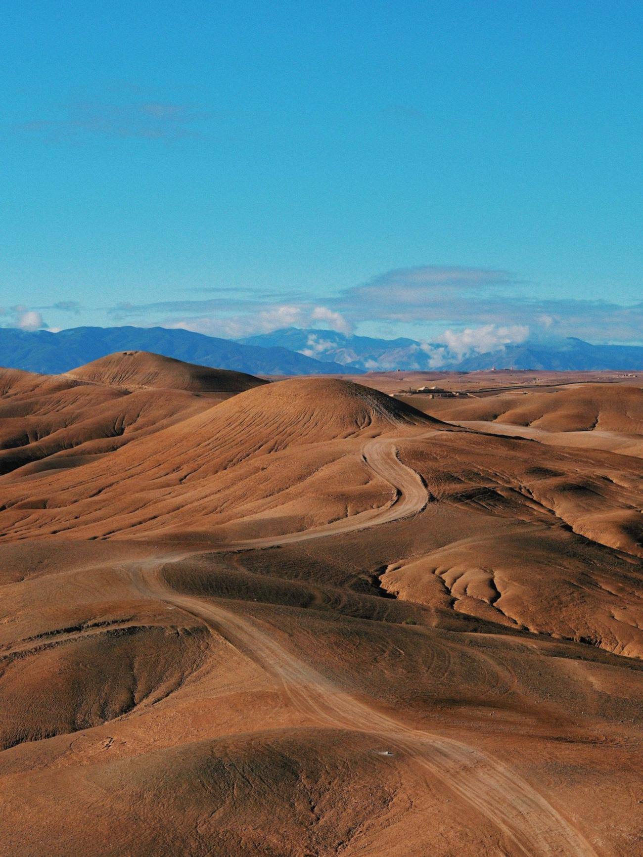 Bare, rounded brown hills of the Agafay Desert with a dirt track and the Atlas mountains under a blue sky