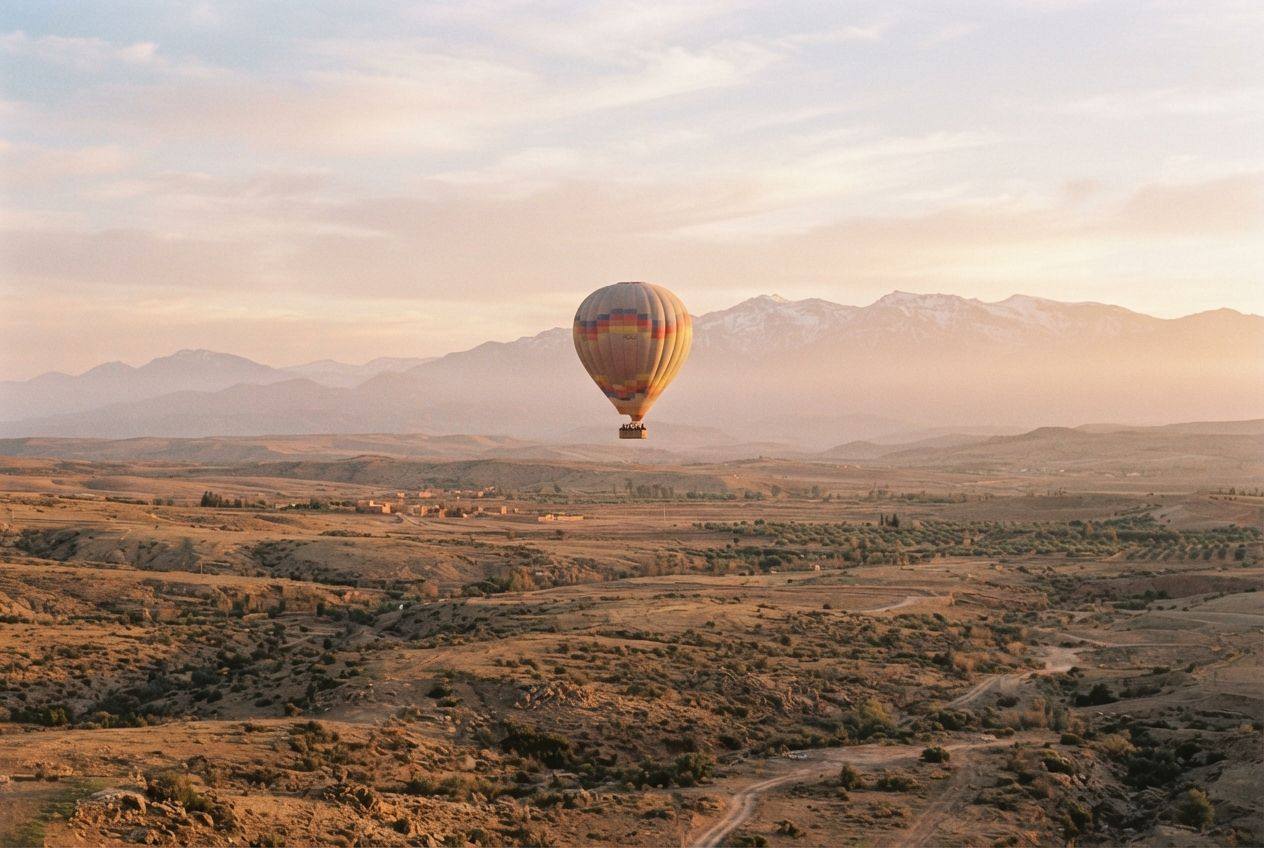 A hot air balloon over dry hills and farmland at sunrise, with the snow-capped Atlas mountains behind